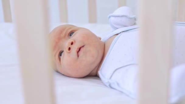 Un pequeño bebé recién nacido yace boca arriba en una cuna sobre una cama de algodón con los ojos abiertos detrás del lado de la cuna — Vídeos de Stock