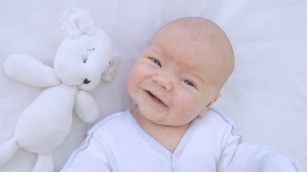 Small newborn baby boy smiles in a cot on a cotton bed with a toy, close-up — Stock Video