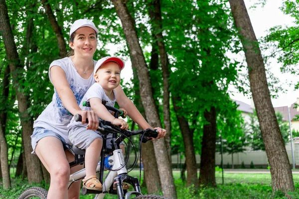 Mãe e filho montam uma bicicleta, mãe carrega uma criança em uma cadeira infantil em uma bicicleta no parque no verão — Fotografia de Stock