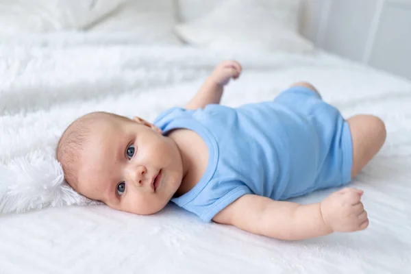 Schattig baby jongen drie maanden oud in een blauw lichaam pak op een wit bed thuis — Stockfoto