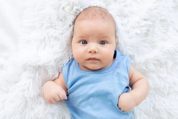 Retrato de un lindo bebé de tres meses de edad en un traje de cuerpo azul en una cama blanca en casa —  Fotos de Stock