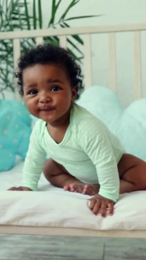 Komik gülümseyen erkek bebek, Amerikan-Afrikalı yatakta mavi pamuklu bir yatakta yeşil bir vücut giysisiyle, dikey video oynuyor.