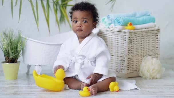 Bebé niño americano-africano juega con un pato de goma en un abrigo blanco después de bañarse cerca del baño, el concepto de higiene y cuidado infantil — Vídeos de Stock