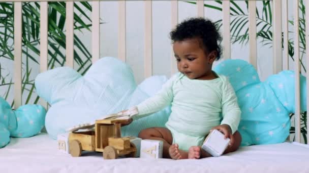 Bebé niño afroamericano juega en la cama con juguetes de madera en un body verde en una cama de algodón azul — Vídeos de Stock