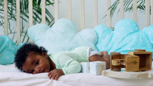Малыш Американо-Африканский милый лежа на кровати с деревянными игрушками в зеленом телесном костюме на голубой хлопковой кровати — стоковое видео