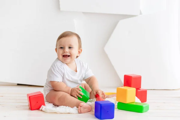 Bebê pequeno feliz seis meses de idade em uma camiseta branca e fraldas brincando em casa em um tapete em um quarto brilhante com cubos coloridos brilhantes — Fotografia de Stock