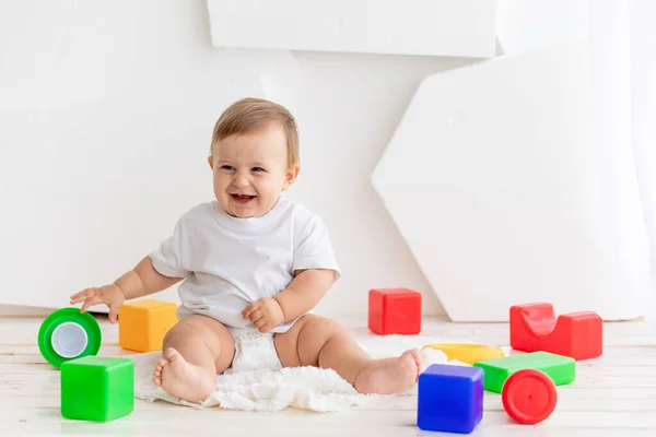Bebê pequeno feliz seis meses de idade em uma camiseta branca e fraldas brincando em casa em um tapete em um quarto brilhante com cubos coloridos brilhantes — Fotografia de Stock