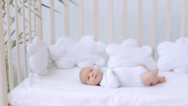 A small newborn baby boy is lying on his back in a cot on a cotton bed with his eyes open in a thoughtful way — Stock Video