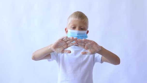 En pojke i en medicinsk skyddsmask tvättar händerna med tvål och skum på en vit isolerad bakgrund, utrymme för text — Stockvideo
