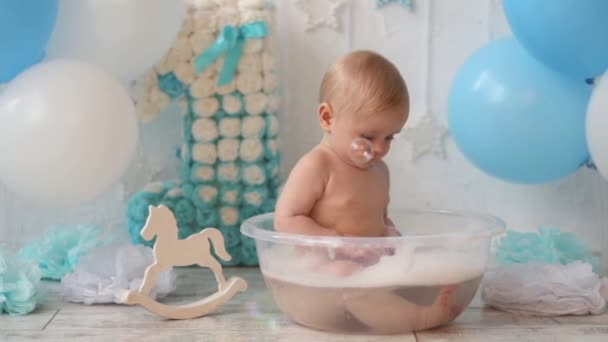 Um menino toma banho em uma bacia com espuma e bolhas de sabão com balões e o número um com uma decoração festiva para o seu aniversário — Vídeo de Stock