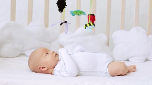 A newborn child plays or looks at toys on a mobile phone in a crib on a white cotton bed at home — Stock Video