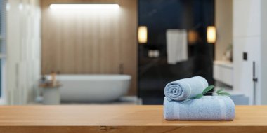 Yumuşak, açık mavi havlular, modern banyo içlerindeki ahşap tezgahın üzerine güzelce katlanmış ve arka planda bulanık bir küvet ve sıcak bir aydınlatma var.