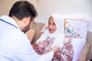 Yaşlı kadın ve doktoru evdeki sağlık kontrolünde terapi detayları ve ilaç dozajı hakkında konuşuyorlar.