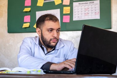 Beyaz üniformalı erkek doktor defter ve dizüstü bilgisayarla kitabı üzerinde çalışıyor.