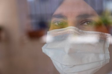 Karantina sırasında pencereden dışarı bakan ve maske takan Arap bir kadın.