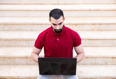 Arap adam dışarıda dizüstü bilgisayar kullanıyor ve merdivenlerde oturuyor.