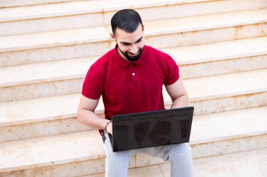 Arap adam dışarıda dizüstü bilgisayar kullanıyor ve merdivenlerde oturuyor.