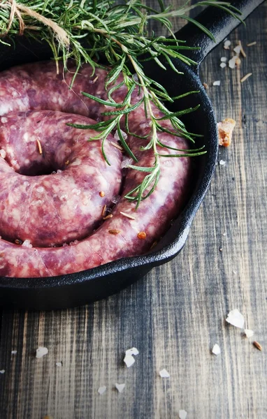 Salsichas de carne crua em uma panela de ferro fundido com alecrim e especiarias, mesa de madeira, foco seletivo — Fotografia de Stock