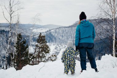 Annesinin elini tutan bir çocuk bir kar yığınında bir dağda duruyor, dağlarda bir aile, bir kış gezisi, arkadan bir manzara, çocuklarla parkta bir yürüyüş.