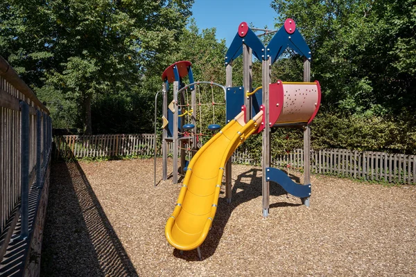 Colorful climbing frame with yellow slide on a small fenced playground with wood chips fall protection