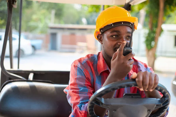 A black engineer or a forklift driver in a warehouse wearing a yellow helmet is talking on the walkie-talkie while sitting in the car.