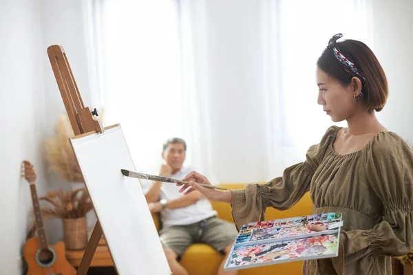 Asian woman who loves art or a female artist painter is holding a paint palette and paintbrushes while happily painting a portrait on white paper at studio.