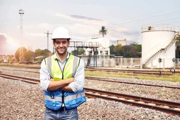 A handsome caucasian Railway engineer or Rail transport technician, wearing a reflective vest and helmet, stands with his arms folded on an outdoor train track.
