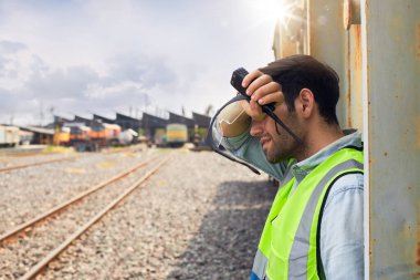 Yeşil güvenlik yeleği giyen bir demiryolu mühendisi ya da demiryolu teknisyeni sıcak ve güneşli bir günde bir yük treninin yanında dinlenmek ya da dışarıda çalışmak için ayakta duruyor..
