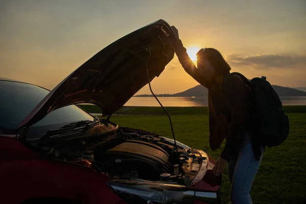 Woman traveler opens the hood car on the front to see the engine that does not start. Young female traveling with a backpack is frustrated by the breakdown of the car in the twilight.