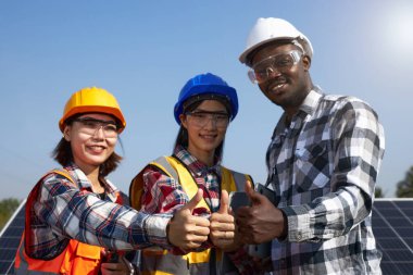 Açık havada birlik göstermek için kask ve güvenlik yelekleri giyen etnik çeşitliliğe sahip bir grup elektrik mühendisi başparmaklarını kaldırdı. Güneş Enerji Yönetimi ve Alternatif Enerji, Temiz Enerji.