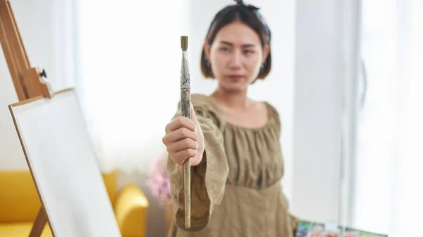 Creative influencer or female blogger an online streaming painting demonstration. A young woman Asian artist holding a paint palette and paint brushes is broadcasting live with a smartphone at home.