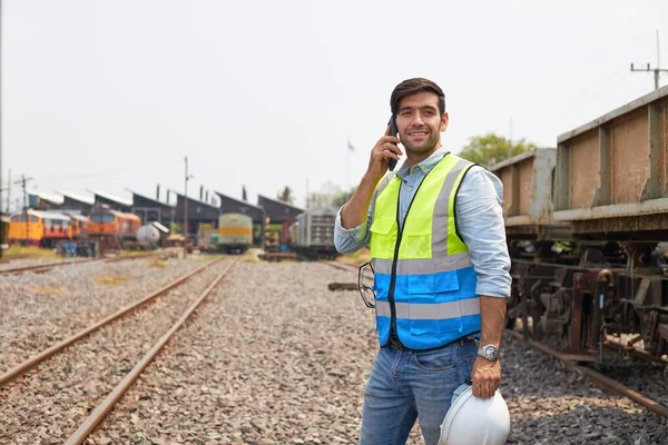 A railroad engineer or maintenance technician in protective clothing is relaxing during the break by smilingly on the phone near an old freight train.