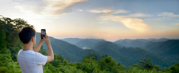 男の観光客は 太陽が彼の電話で下に行くと 夕方には山の風景の写真を撮る アジアの男性は 週末や雨季に自然旅行を楽しむ — ストック写真