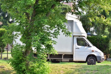 Ağaçların arkasında beyaz bir teslimat kamyonu var. Yaz.