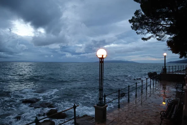 Promenade with lights on and the sea in the early morning before sunrise, Opatija, Croatia. Scenic view of stormy sea, sky in clouds and bright lanterns on rainy autumn morning