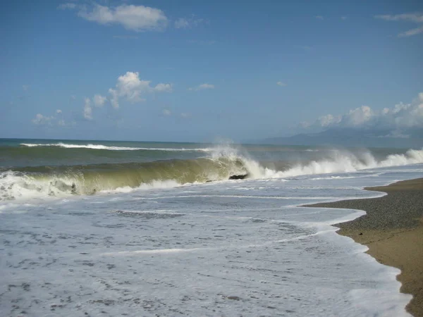 Seascape stormy sea on the coast of Calabria on a sunny summer day. Storm on the Tyrrhenian Sea, wave and sea foam on a sandy beach against a background of blue sky and mountains on the horizon