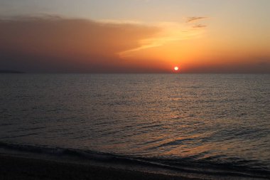 Sahilde gün batımı manzarası, sıcak bir sonbahar akşamı çakıl taşı sahilinde turuncu bulutlarla ifade edilen gökyüzü. Güzel gri ve turuncu bulutlar, parlak güneş diski ufukta ve sakin deniz, Calabria, İtalya