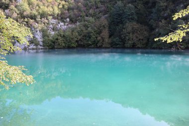Güneşli bir günde, Hırvatistan 'ın Plitvice Gölleri Ulusal Parkı' nda, sonbahar yapraklı kayalar ve ağaçlarla çevrili, su yüzeyinin üzerinde turkuaz su ve sisli resim gölü