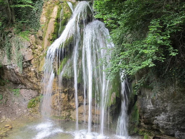 Kırım 'ın yeşil yosunlarıyla kaplanmış dik kayalarla kaplı bir vadideki resim şelalesi Dzhur-Dzhur. Şelalenin manzarası kayaların arka planında güçlü su fıskiyeleri, parlak yeşil yosunlar, yaz günü yeşil ağaç yaprakları, Kırım.