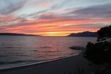 Hırvatistan 'da deniz gün batımında kıyıdaki adalar ve dağlar görülüyor. Kırmızı-turuncu bulutlar, ufukta Brac adası, kumsalda bir yaz akşamında parlak gökyüzü ve deniz.