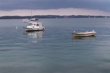 Gök gürültülü gri bulutlar, ufukta yağmur yağarken sakin deniz martıları yağmurlu bir günde beyaz bir teknede oturuyor, Hırvatistan. Deniz manzaralı martılar, demir atmış tekneler ve ufukta gök gürültülü bulutlar, denizin sakin sularına yansıyan bulutlar.