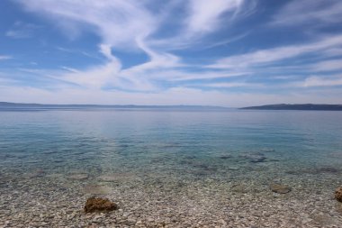 Hırvatistan 'ın güneşli bir yaz gününde mavi gökyüzü ve turkuaz şeffaf deniz. Hırvatistan kıyılarında manzaralı deniz manzarası, altında çakıl taşlarının göründüğü açık mavi deniz suyu, ufukta Brac adası.