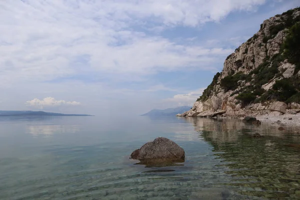 Seascape calm at sea in a picturesque bay on the sea coast of Croatia on a summer day. Calm clear water of the Adriatic Sea, in which clouds are reflected and stones are visible at the bottom against the background of high rocks on the shore