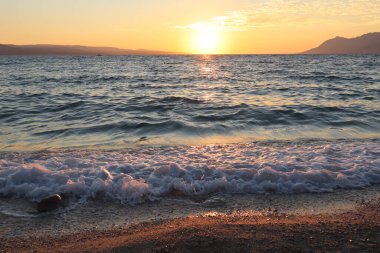 Sahilde gün batımı manzarası, Hırvatistan 'ın su yüzeyinde güneş parlaması ve ıslak kum. Kumsaldaki fırtınalı deniz ve çakıl taşları bir sonbahar akşamında batan güneşin ışınlarıyla aydınlatılır.