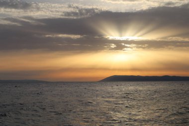 Ada siluetiyle deniz üzerinde manzaralı bir gün batımı. Hırvatistan kıyılarında gün batımında gökyüzündeki bulutların arasından güneş ışıldıyor