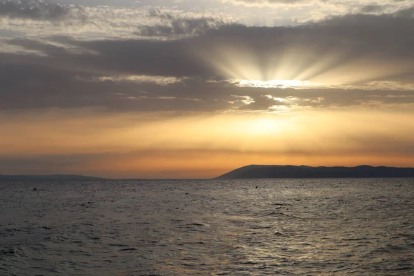 Ada siluetiyle deniz üzerinde manzaralı bir gün batımı. Hırvatistan kıyılarında gün batımında gökyüzündeki bulutların arasından güneş ışıldıyor