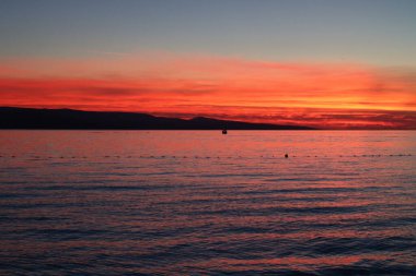 Denizde gün batımı, parlak kırmızı gökyüzü, bulutların ardında güneş ve ufukta ada silueti.