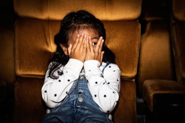 Sevimli küçük kızlar korku ve heyecanla dolu heyecanlı bir sinemada oturup eğlenceli filmler izler. Yüzlerini örtmek için elleri kullanır..