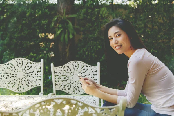 日陰の庭で朝リラックスした白い合金テーブルの上に座っているアジアの女性 彼女の日常生活は彼女の暇な時間に快適に座るために彼女の携帯電話にあります — ストック写真