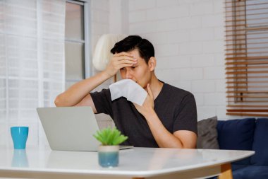 Yakışıklı Asyalı adam evde hasta, ateşi, tıkalı burnu, hapşırması, akışkan burnu, elinde tuvalet kağıdı, burnunda sıvı ile oturuyor..